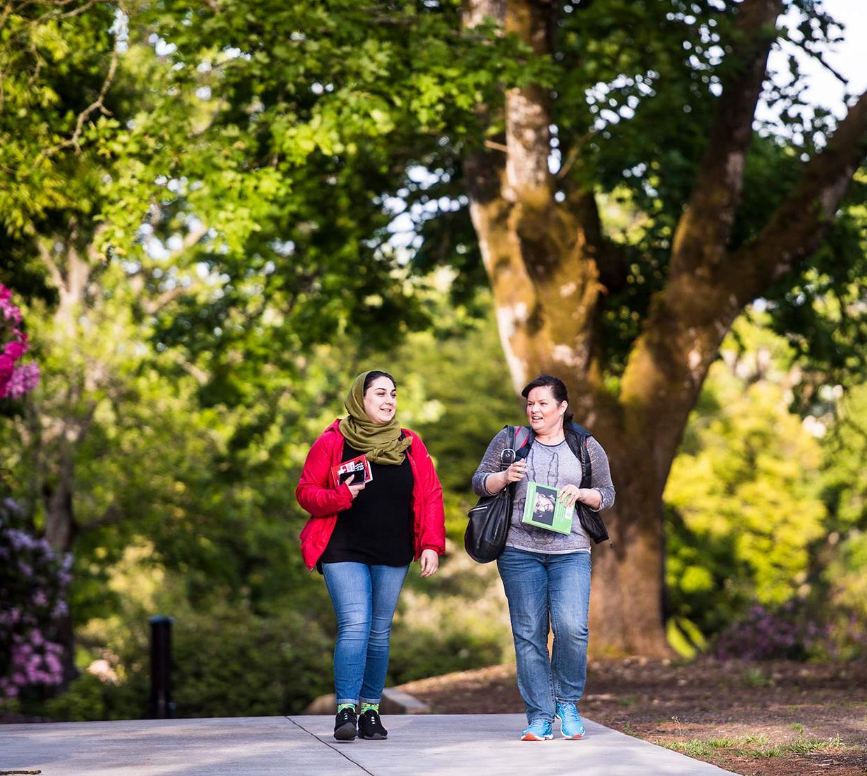 Two female students walking through campus in front of tree